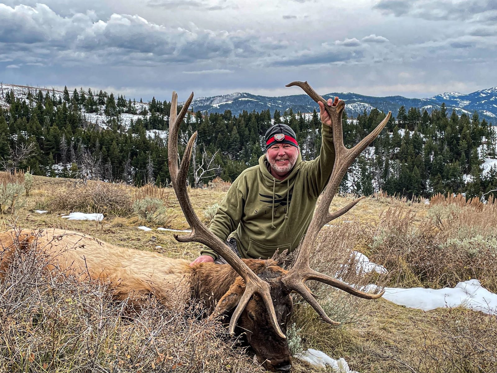 A hunter poses with a harvested elk in a snowy, mountainous landscape.