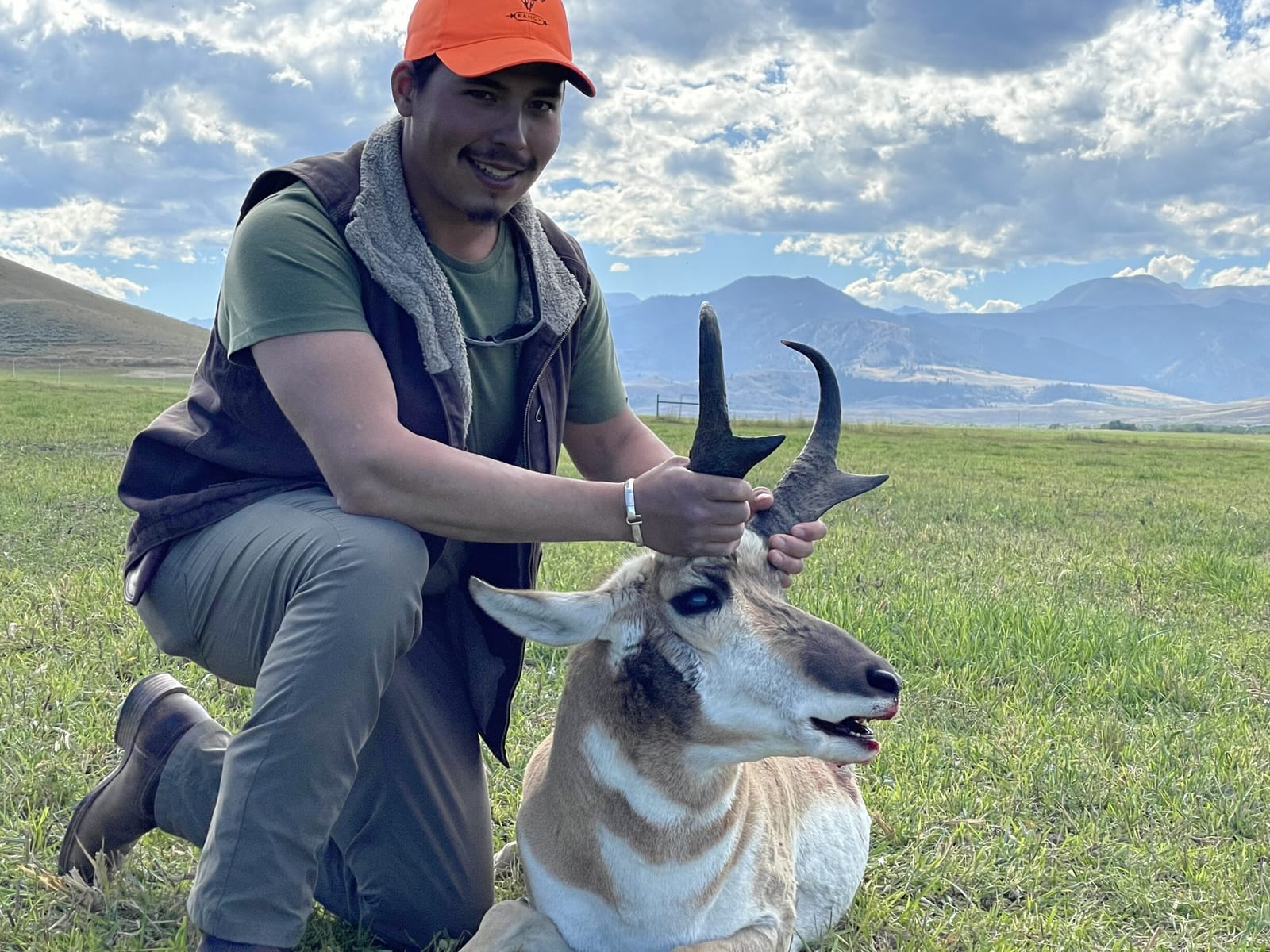 A hunter kneels next to a harvested pronghorn in a grassy field with mountains in the background.