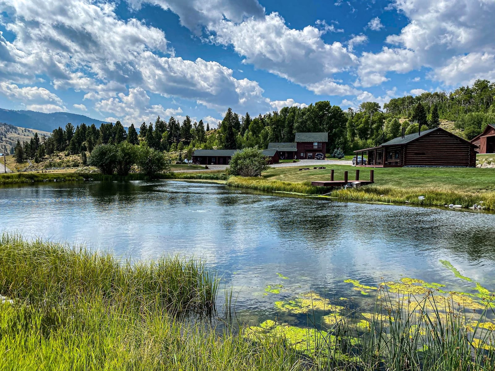 Log cabins are situated by a pond with green grass and trees, backed by mountains under a cloudy sky.
