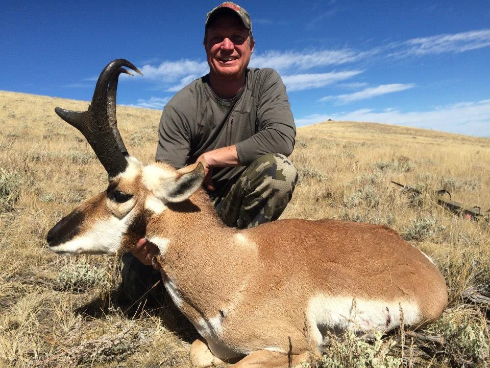 A hunter kneels next to a harvested pronghorn in a grassy field under a blue sky.