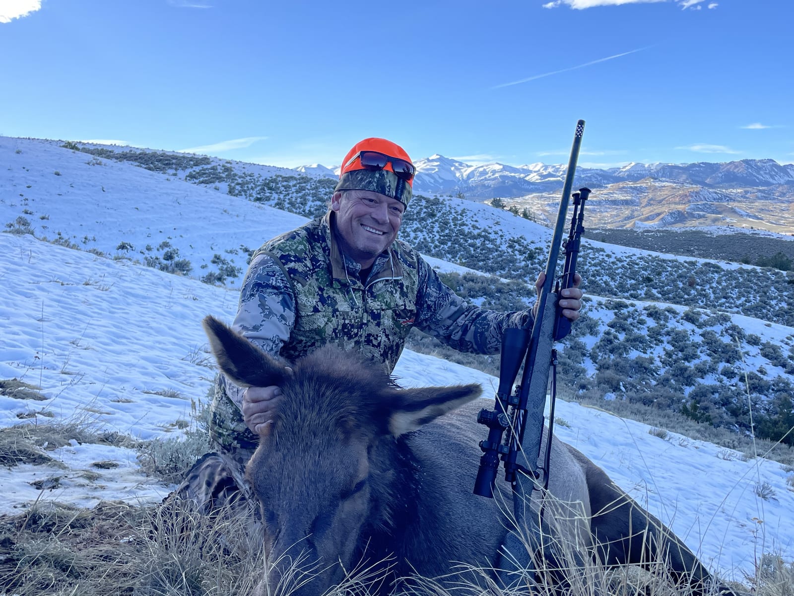 A hunter in camouflage and an orange hat poses with a harvested elk in a snowy, mountainous landscape.