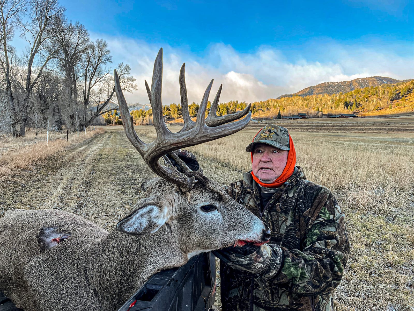 A hunter in camouflage poses with a harvested whitetail deer in a field with trees and mountains in the background.