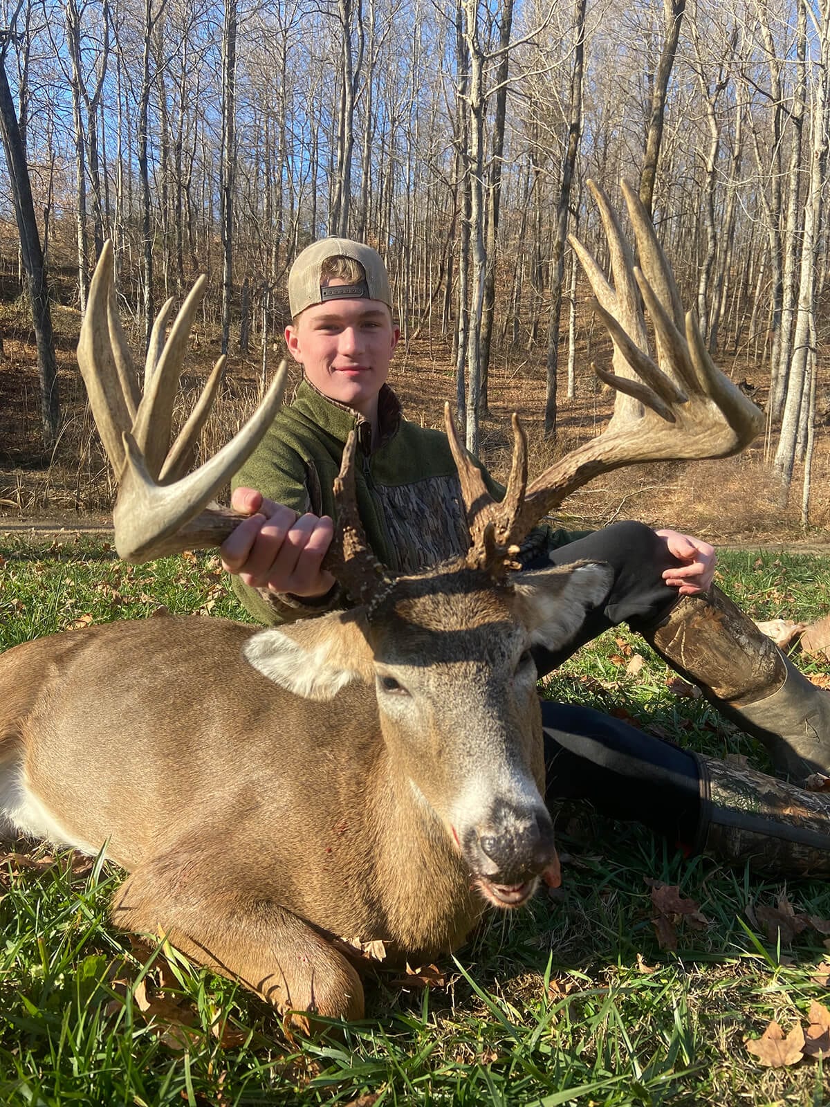 A young man poses with a large harvested whitetail deer in a wooded area.