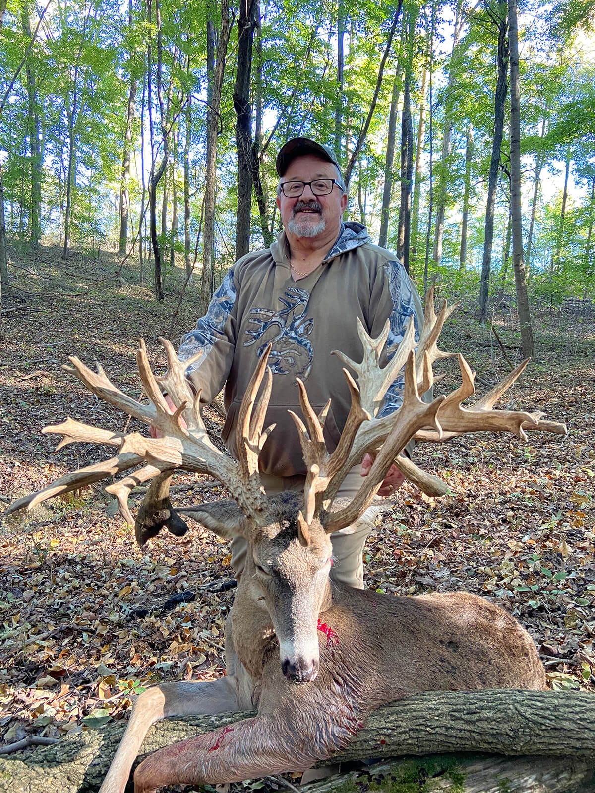 A hunter poses with a large whitetail deer harvest in a wooded area.