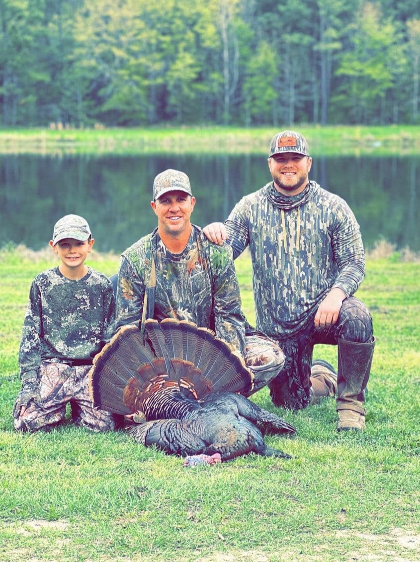 Three hunters, including a child, pose with a harvested wild turkey in front of a pond.