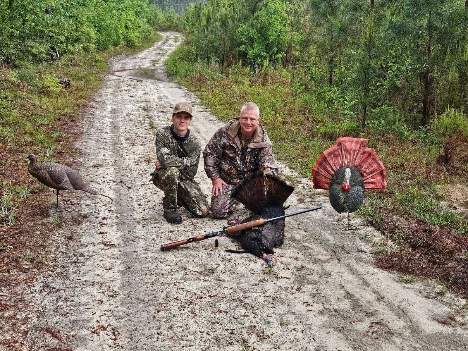 Two hunters pose with a harvested wild turkey and hunting decoys on a dirt road.