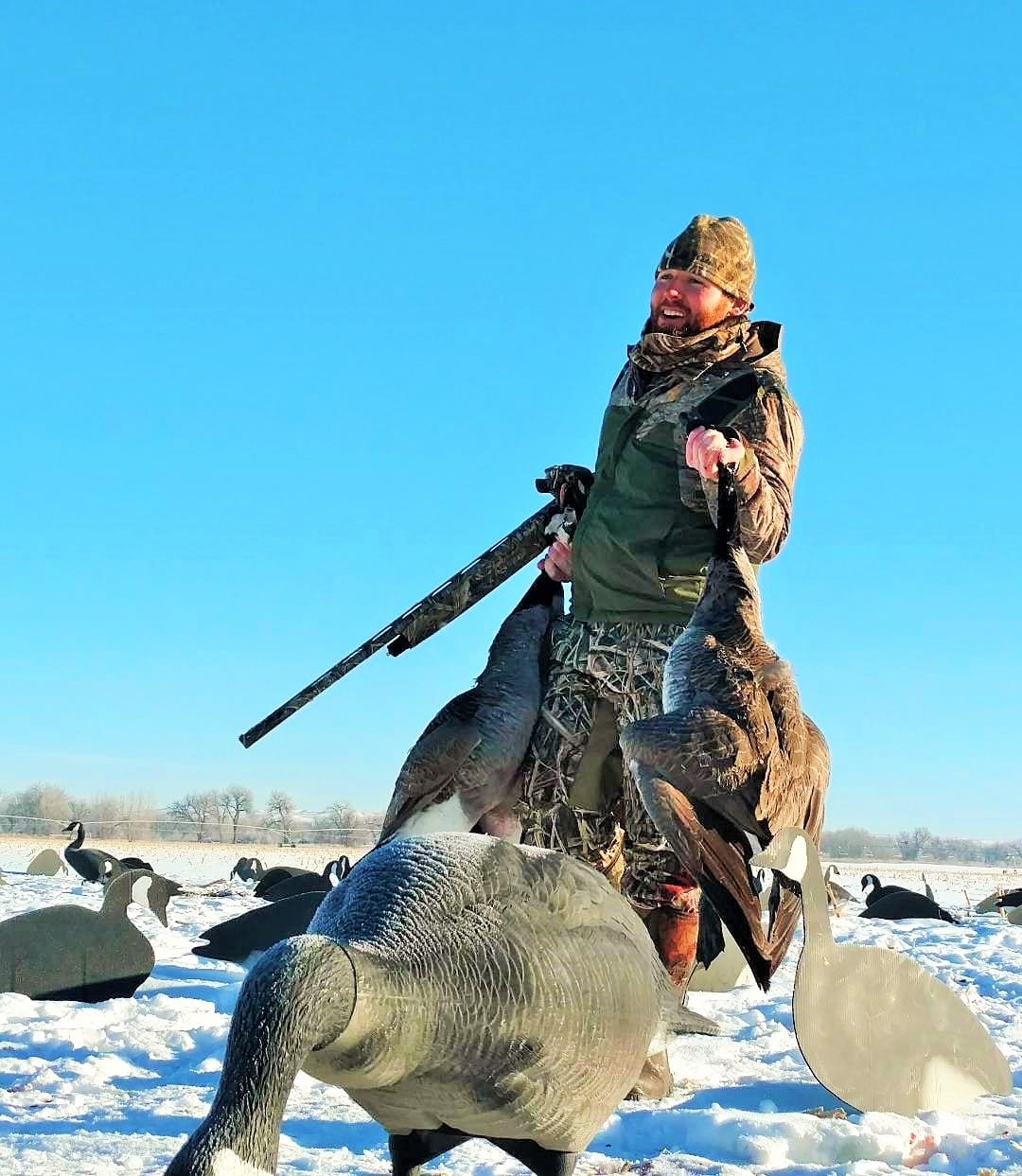 A hunter in camouflage holds two harvested Canada Geese in a snowy field with decoys.