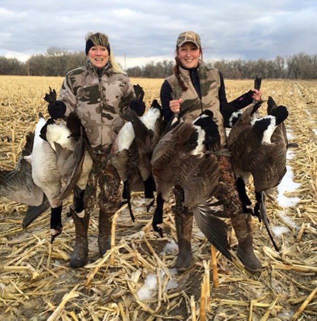 Two women in camouflage stand in a field holding several harvested Canada Geese.
