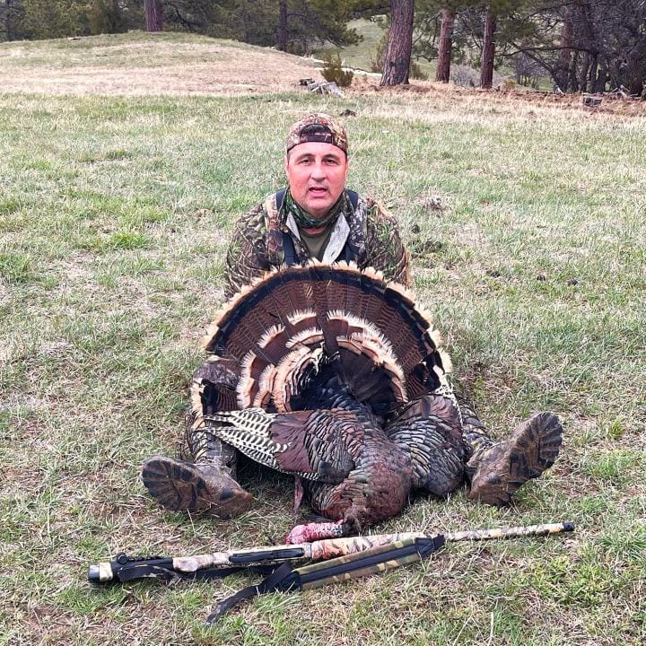 A hunter in camouflage sits on the grass, posing with a harvested wild turkey and a shotgun.