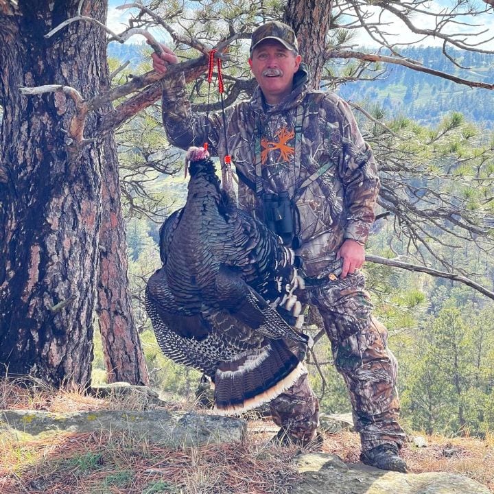 A hunter in camouflage poses with a harvested wild turkey in a wooded area.