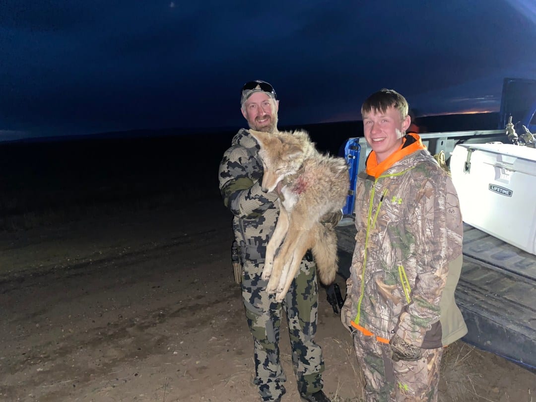 Two hunters pose with a harvested coyote at dusk.