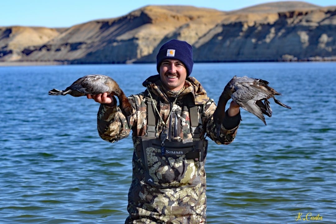 A hunter stands in water holding two harvested redhead ducks with a lake and hills in the background.