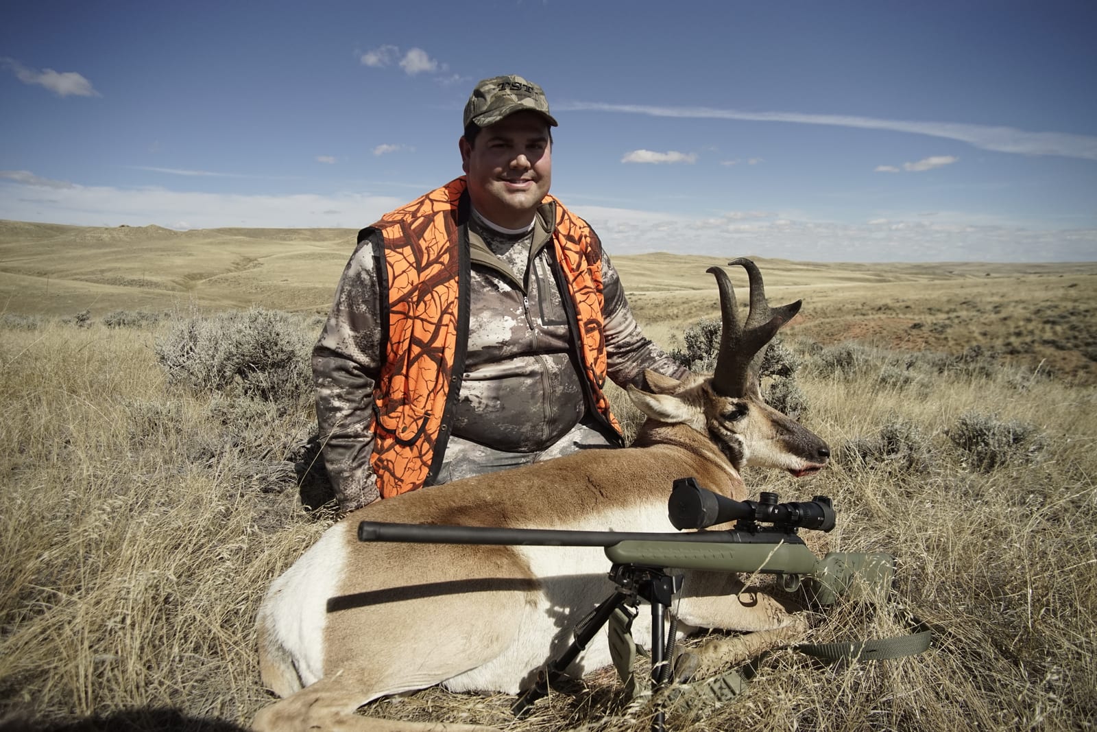 A hunter in camouflage and an orange vest poses with a harvested pronghorn and a rifle in a dry, grassy field under a blue sky.