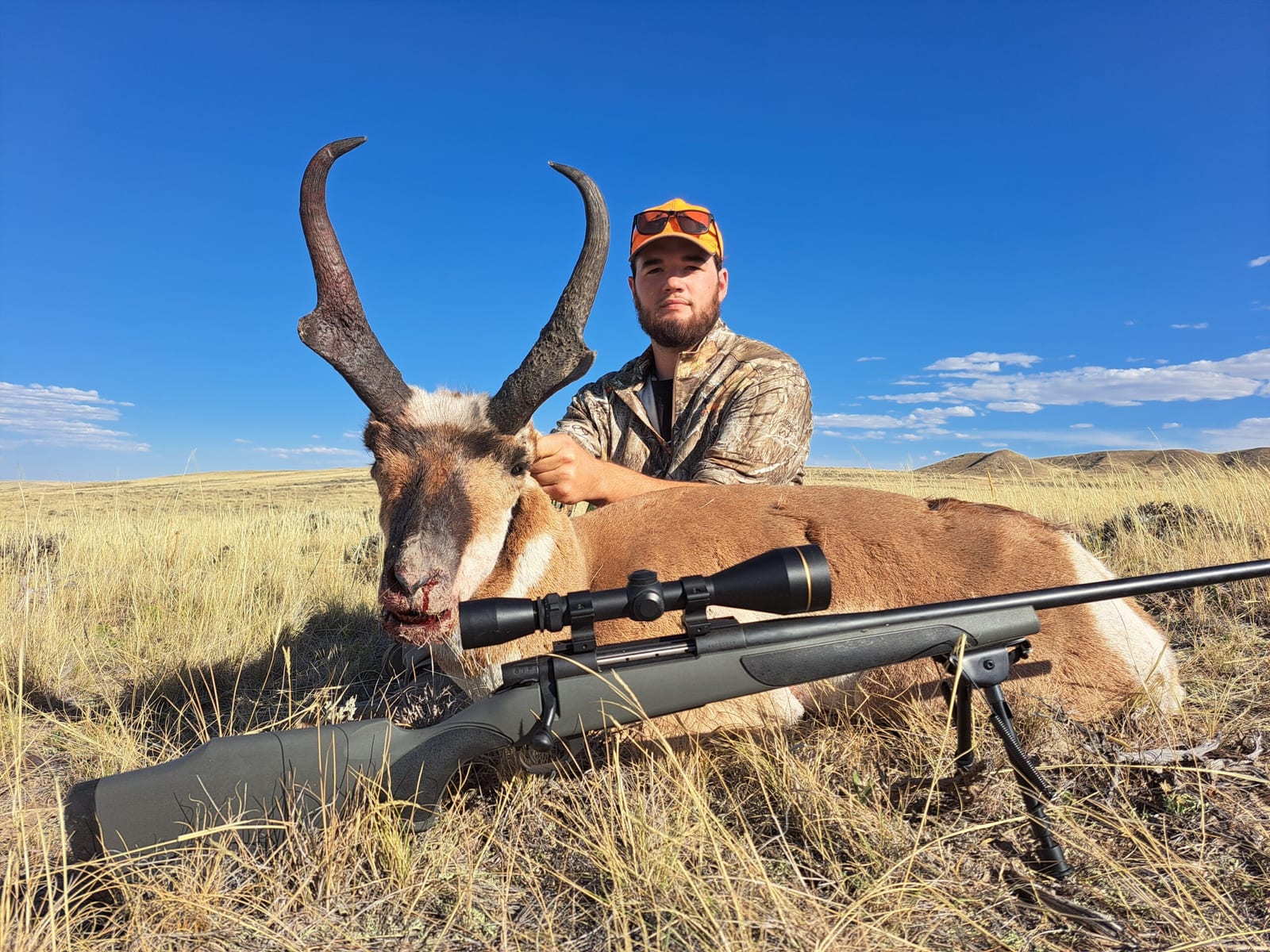 A hunter poses with a harvested pronghorn in a grassy field under a blue sky.