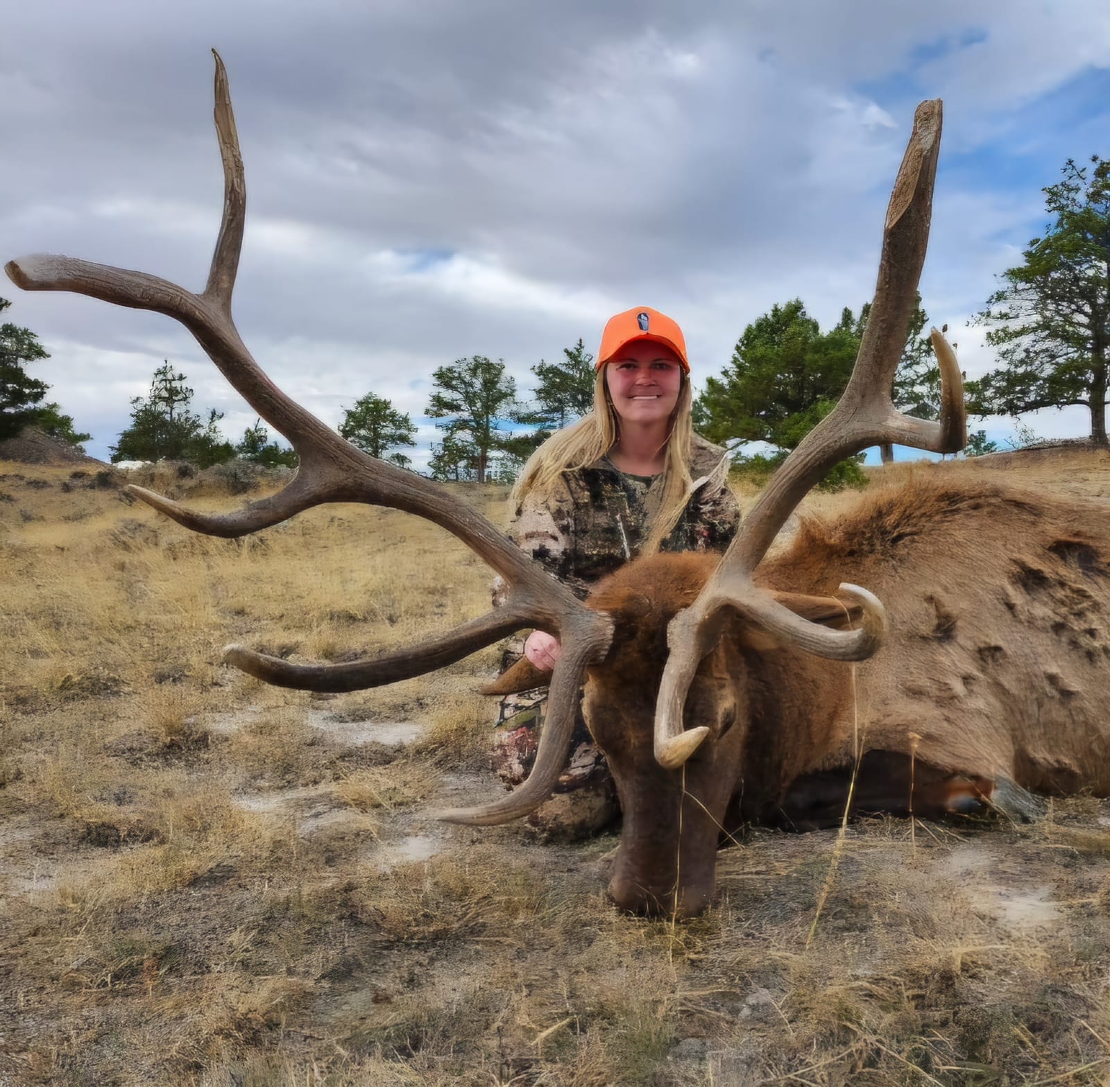 A hunter in camouflage and an orange hat poses with a harvested elk in a grassy field under a cloudy sky.