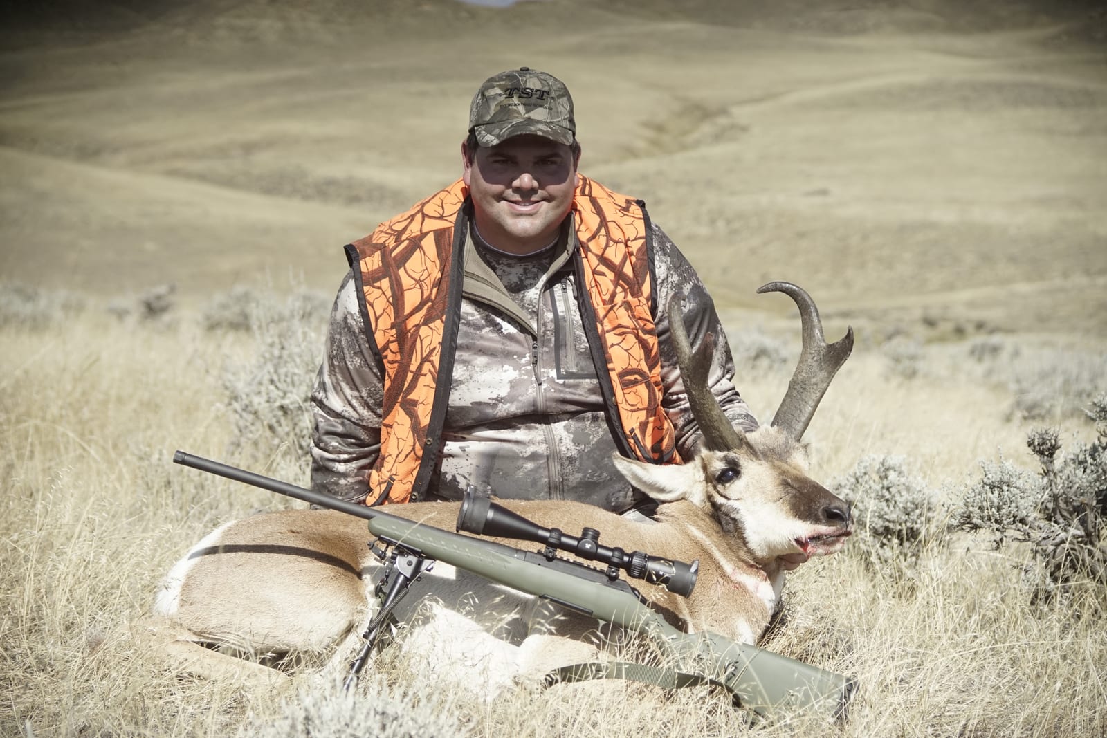 A hunter in camouflage and an orange vest poses with a harvested pronghorn and a rifle in a grassy field.