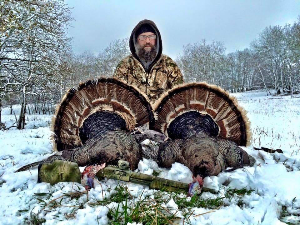 A hunter poses with two harvested wild turkeys in a snowy, wooded landscape.