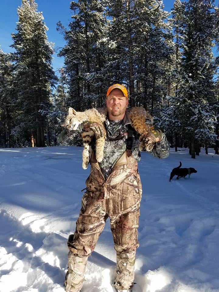 A hunter in camouflage stands in a snowy forest, holding a harvested bobcat and a mountain lion over his shoulders, with a dog in the background.