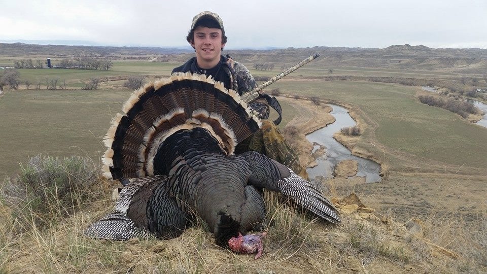 A young hunter poses with a harvested wild turkey on a hillside overlooking a river valley.
