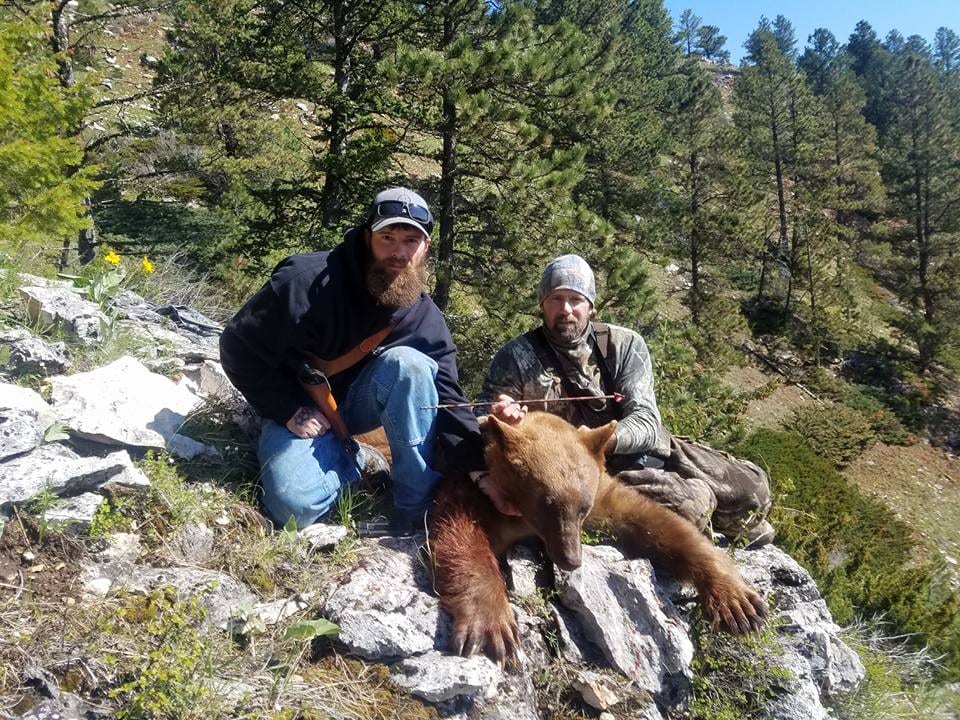 Two hunters pose with a harvested black bear on a rocky, forested hillside.