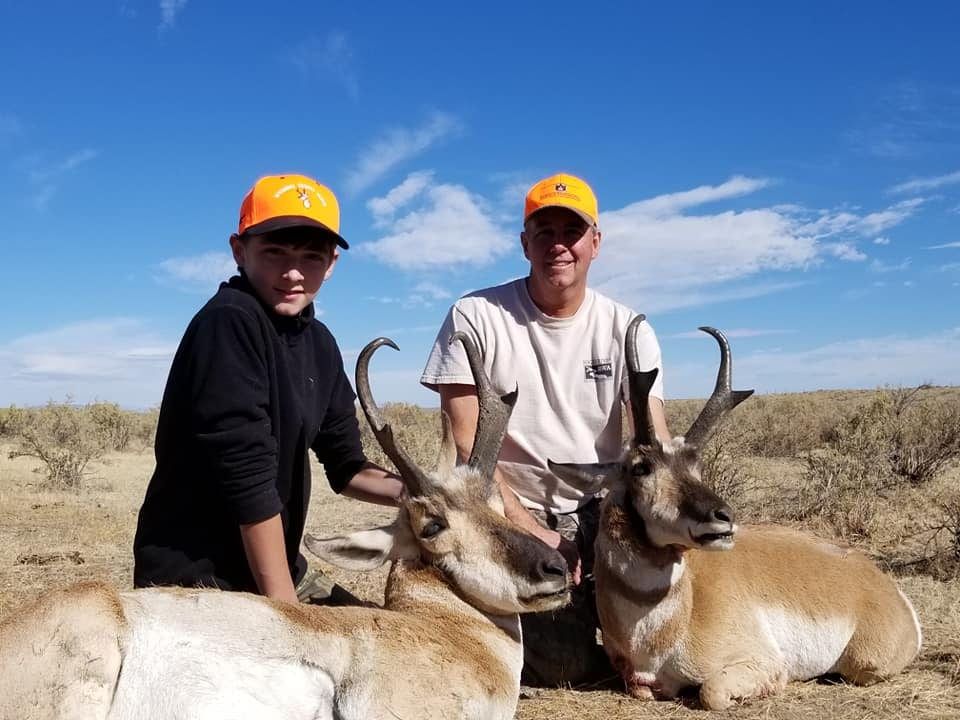 Two hunters pose with two harvested pronghorn antelope under a blue sky.