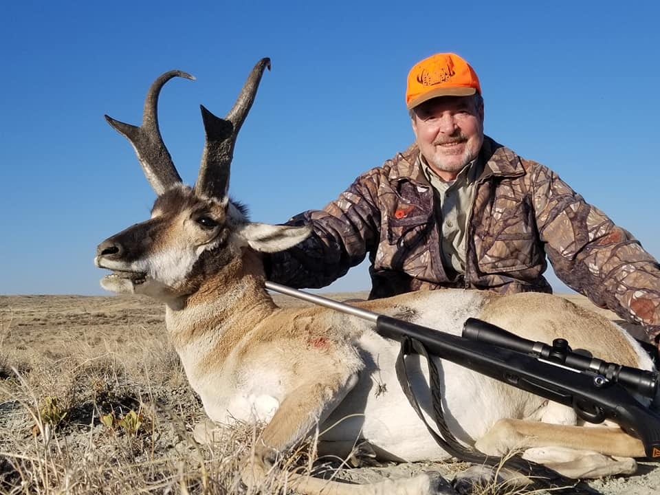 A hunter in camouflage and an orange hat poses with a harvested pronghorn in a dry, grassy field under a clear blue sky.