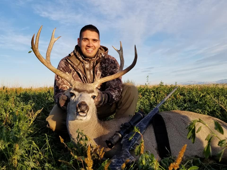 A hunter poses with a harvested mule deer in a field.