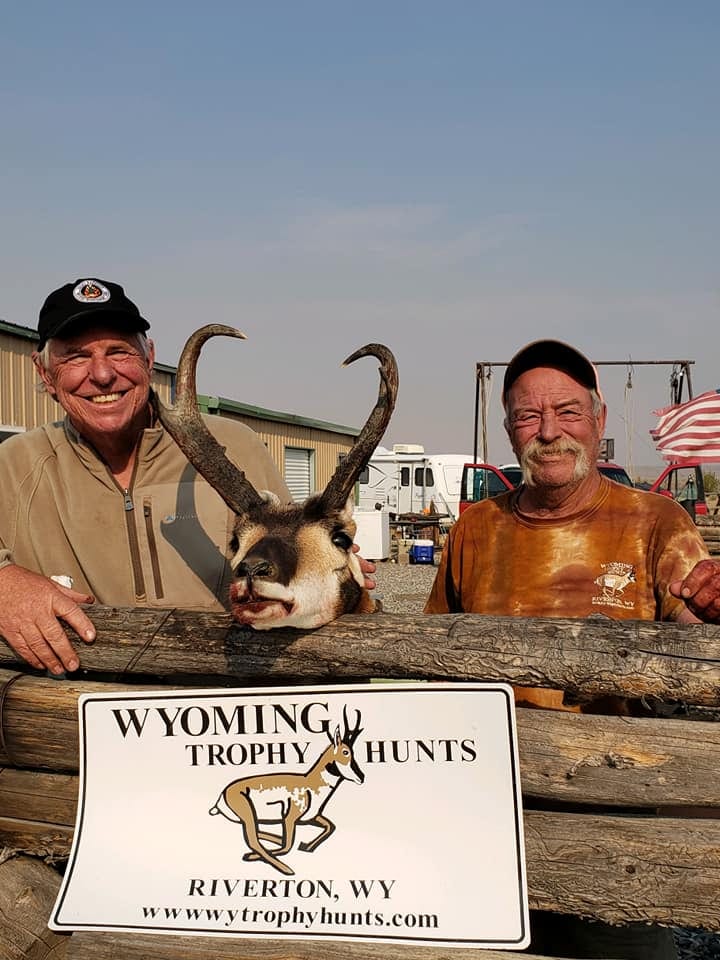 Two men pose with a harvested pronghorn head and a "Wyoming Trophy Hunts" sign.