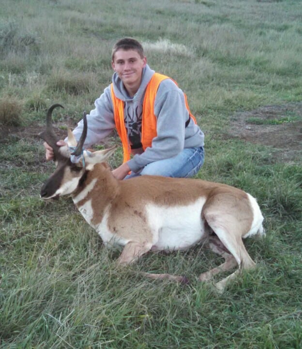 A young hunter kneels next to a harvested pronghorn in a grassy field.