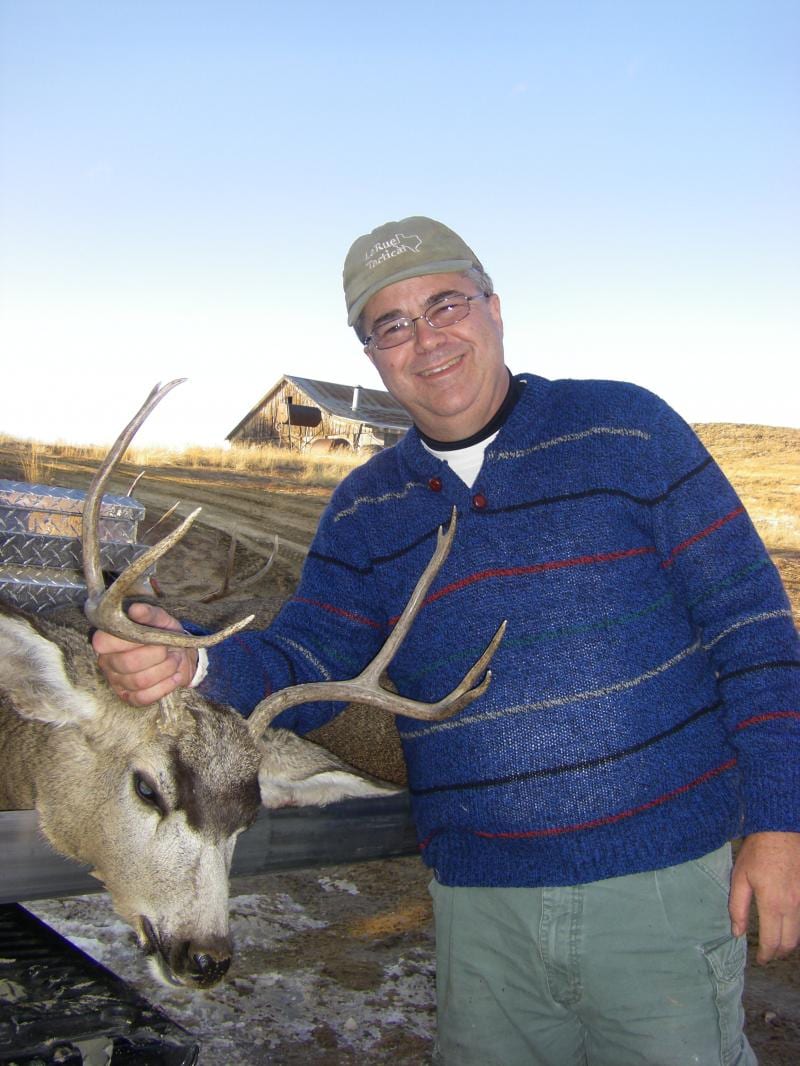A man in a blue sweater and cap poses with a harvested mule deer in a field with a rustic building in the background.