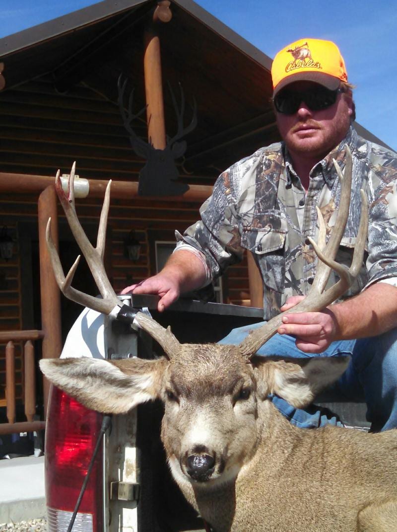 A hunter poses with a harvested mule deer in front of a lodge.