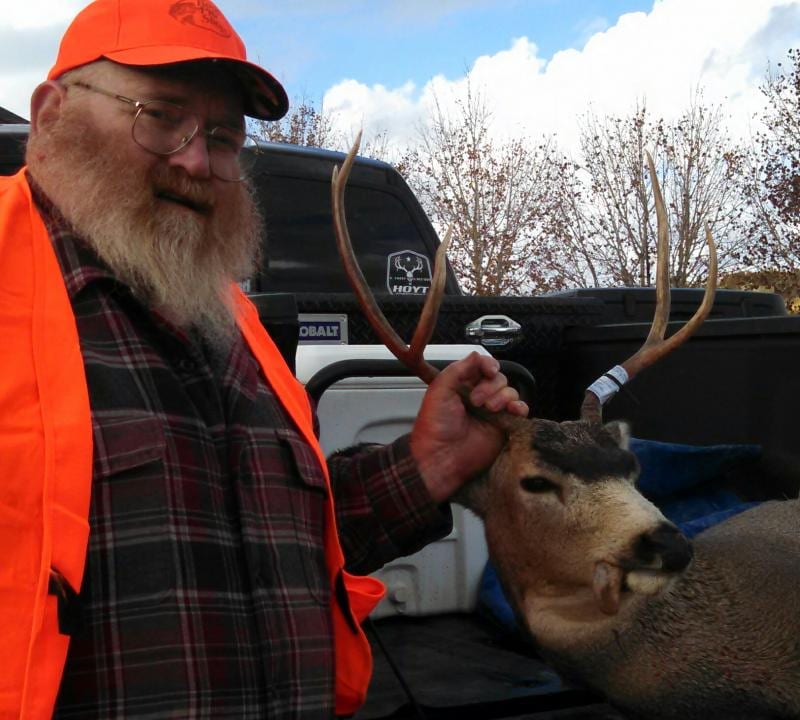 A hunter in an orange vest and hat poses with a harvested mule deer.